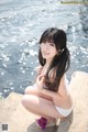 A woman in a white bikini sitting on a rock by the water.