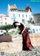 A woman in a red and black dress leaning against a stone wall.