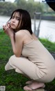 A woman in a beige dress sitting on the grass by the water.